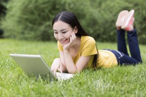 student lying on grass using laptop on campus at college