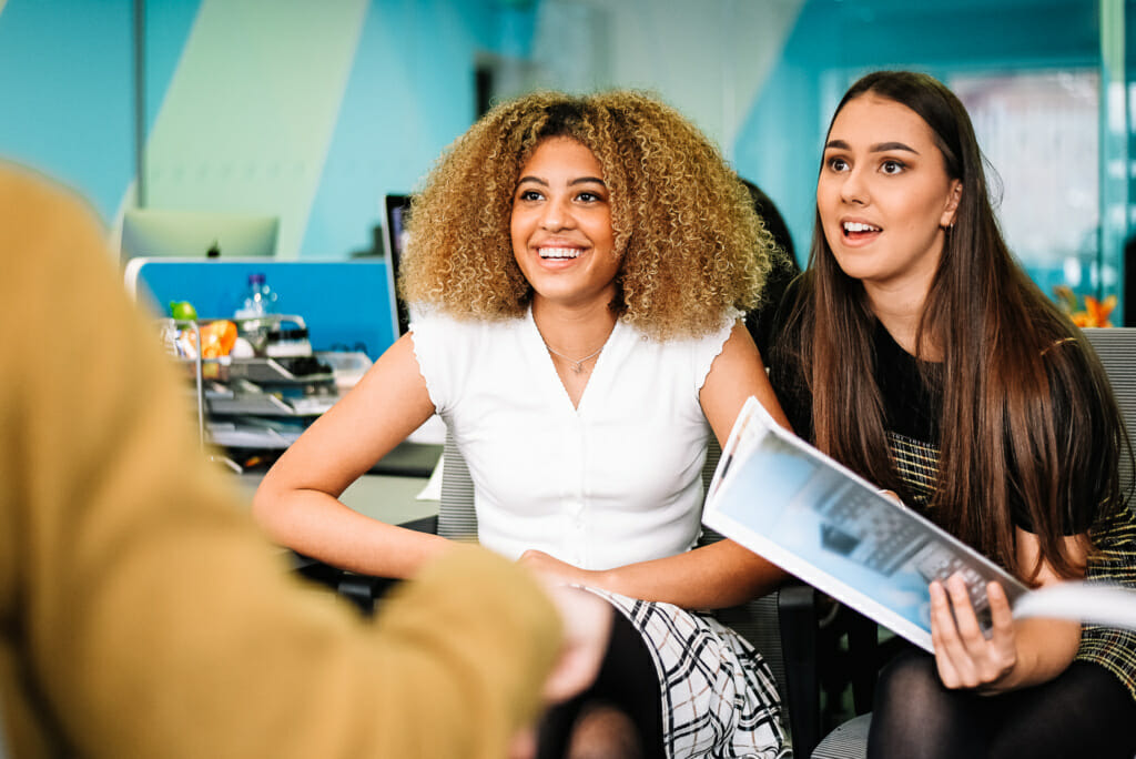 2 females students, one holding a booklet. Both smiling talking to a male.